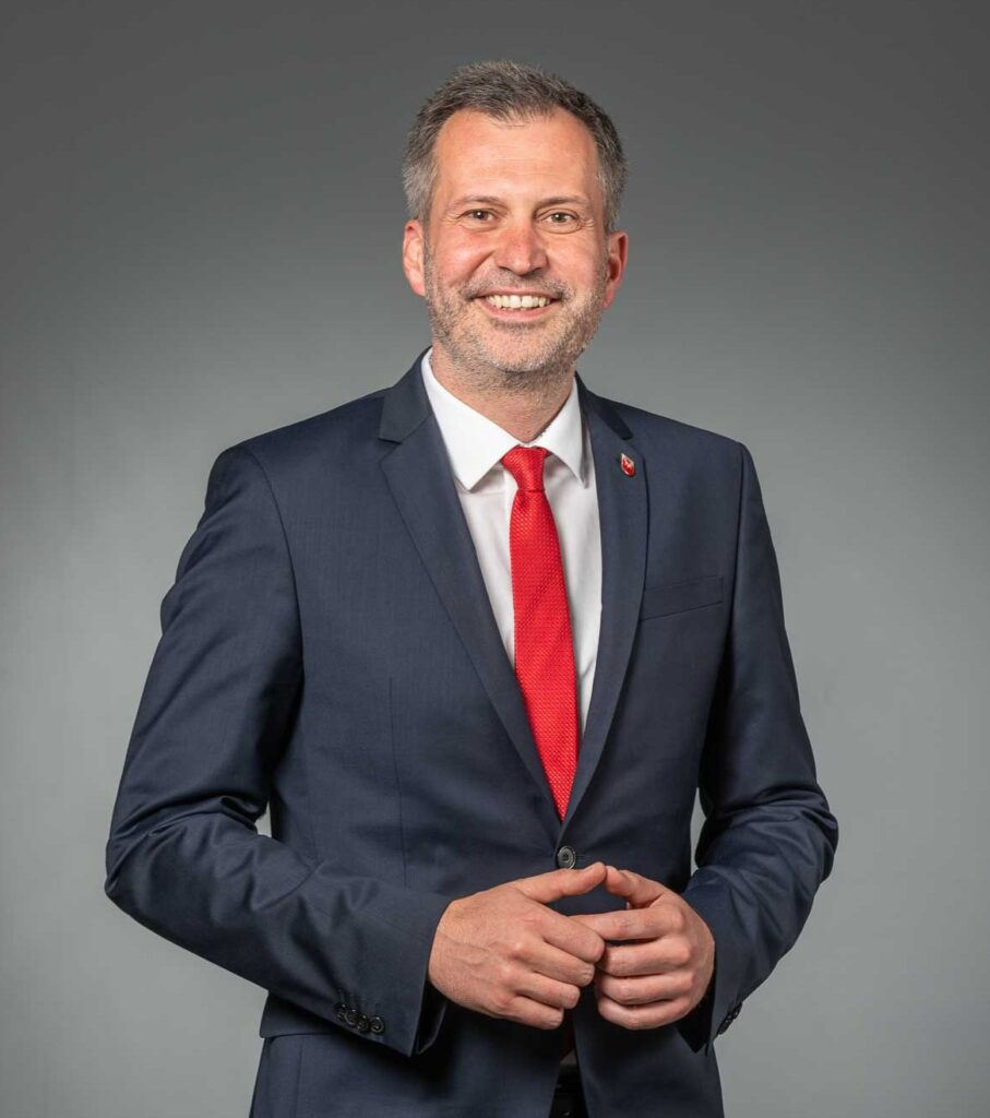 Oberbürgermeister Tobias Schick in einem dunkelblauen Anzug mit roter Krawatte steht mit verschränkten Händen vor einem grauen Hintergrund.